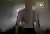 A shirtless man seen from behind at night in mist and city lights, a large circular black-and-grey tattoo across his upper back showing a hand, a crystal and the spokes of a wheel